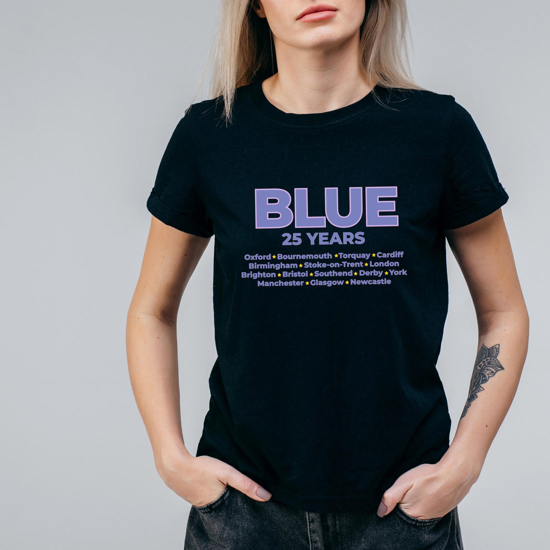 May include: Black t-shirt featuring the word "BLUE" in purple, with "25 YEARS" beneath. A list of UK cities, including Oxford, Birmingham, and Brighton, is printed below. The shirt has a crew neck.